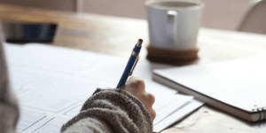 a close up of someone in a sweater writing on paper on top of a table with a mug in the background