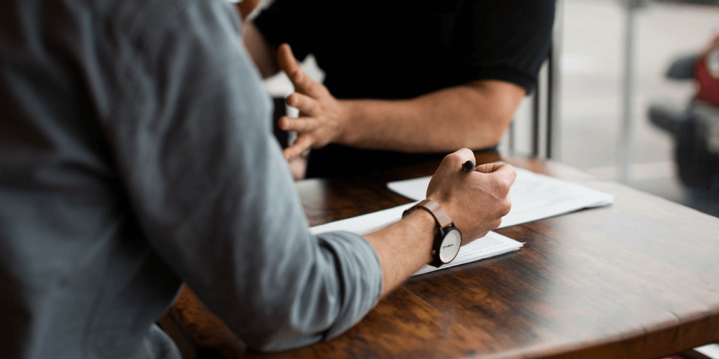Image of two men zoomed in talking at a table, one with a pen in hand ready to sign paperwork