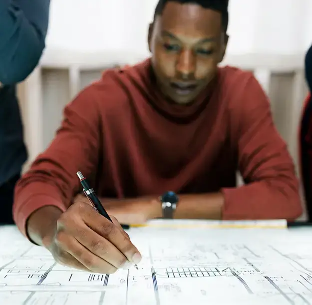Focused professional using a pencil to analyze complex structural plans and documents on a large desk.