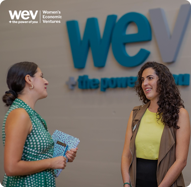 Two women talking in front of the Women’s Economic Ventures logo.